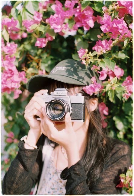 A woman in a garden taking photos with a vintage camera surrounded by pink flowers.