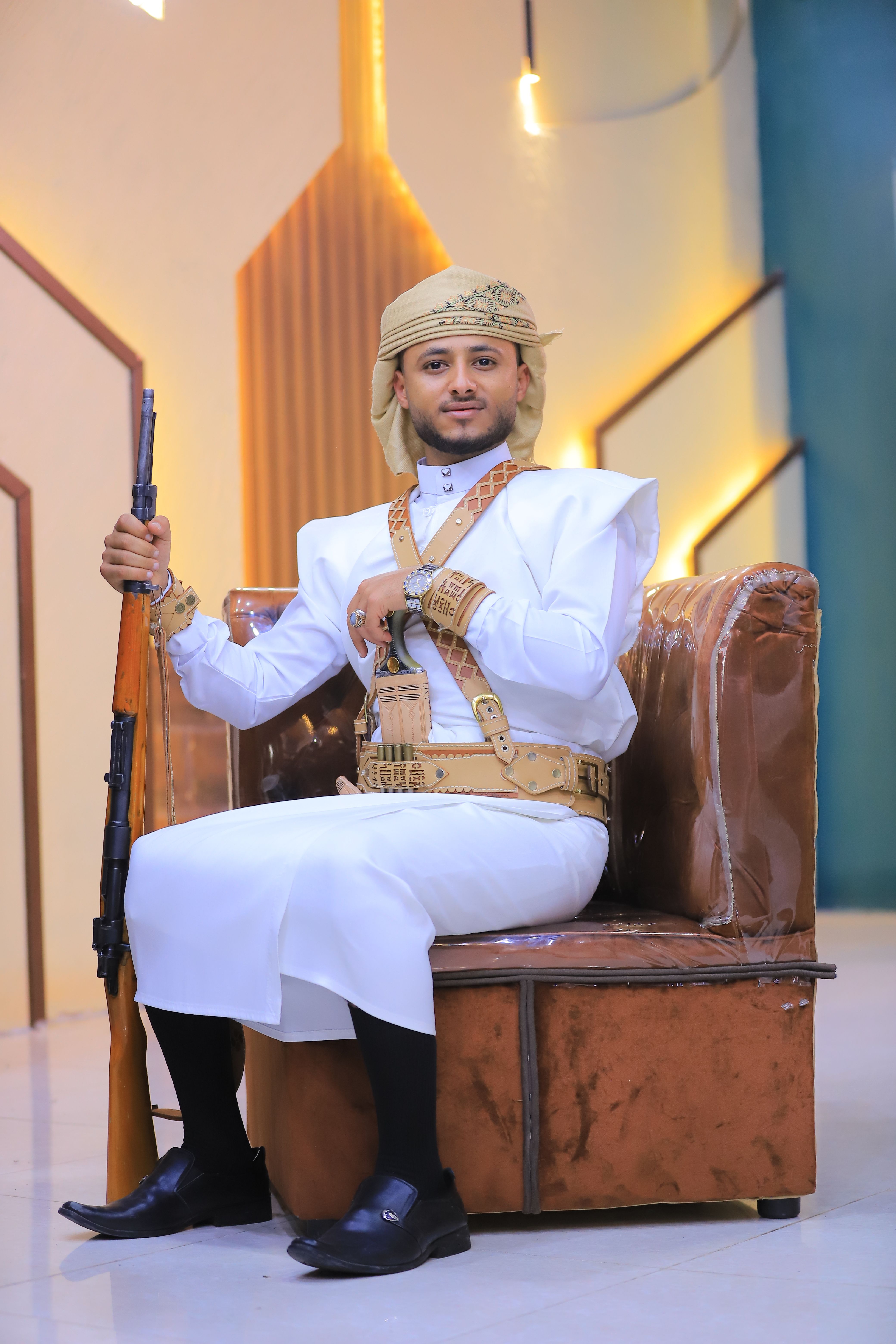 Man in traditional Yemeni clothing holding a rifle indoors with vibrant decor.