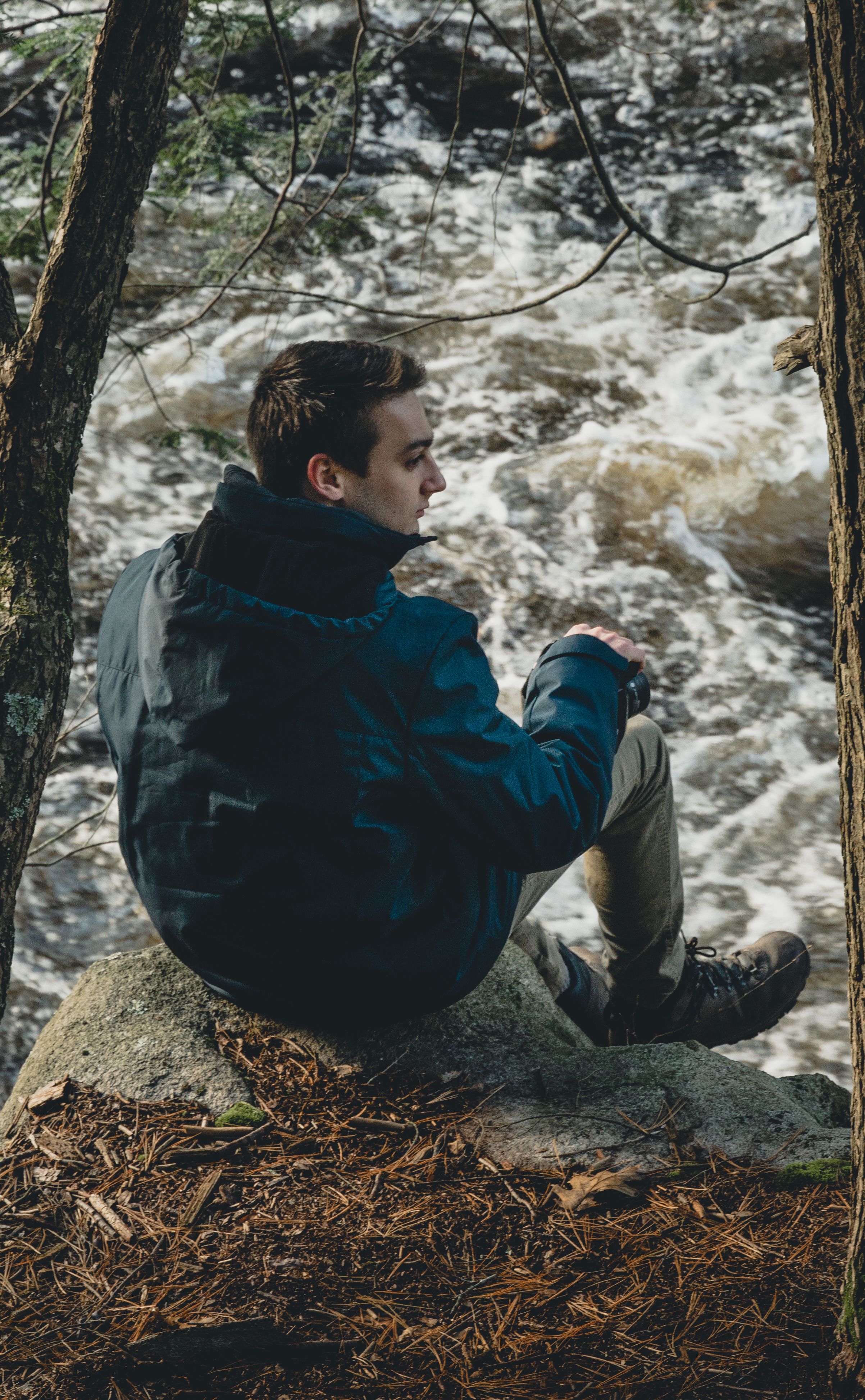 A man in hiking gear sits on a rock by a flowing river surrounded by forest, enjoying the serene nature atmosphere.