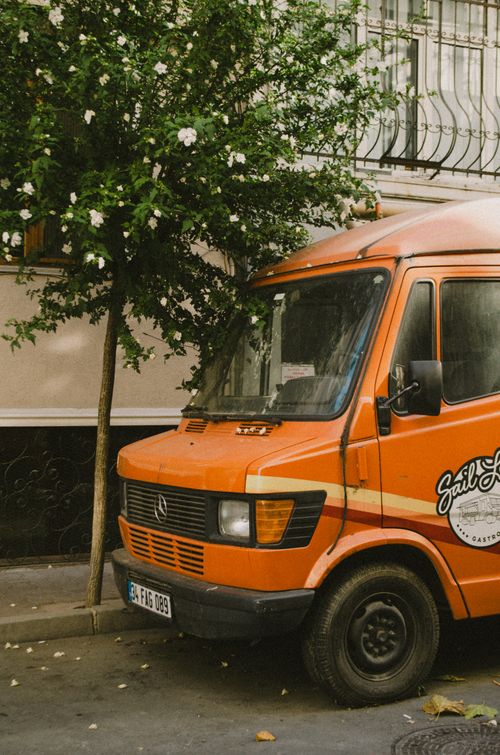 Free A vintage orange van parked in a cozy urban setting beside a flowering tree. Stock Photo