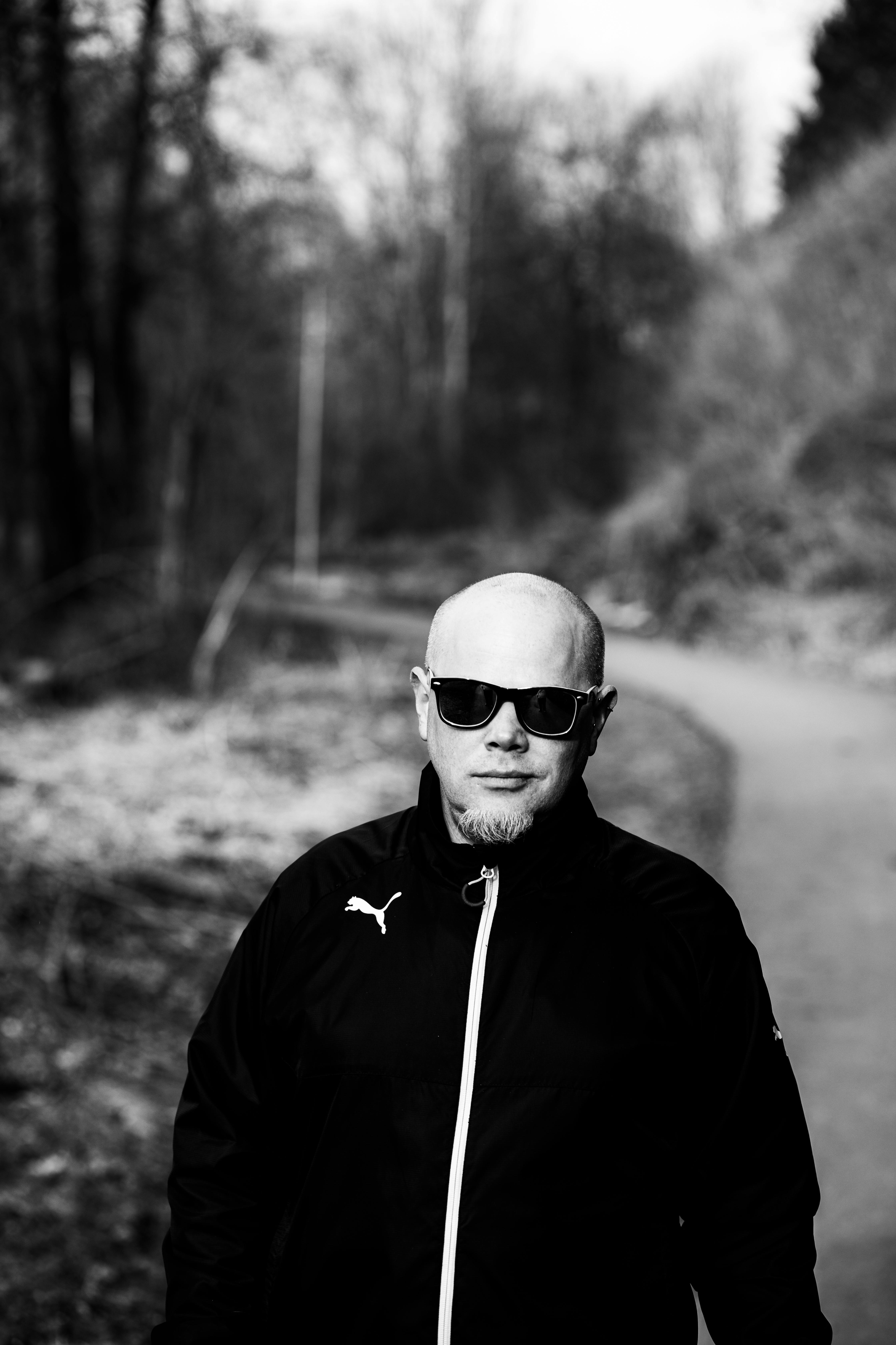 Bald man wearing sunglasses walks in a park, captured in black and white for a dramatic effect.