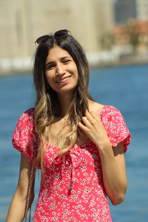Free A smiling young woman in a floral dress stands by a serene waterside, enjoying a sunny summer day. Stock Photo