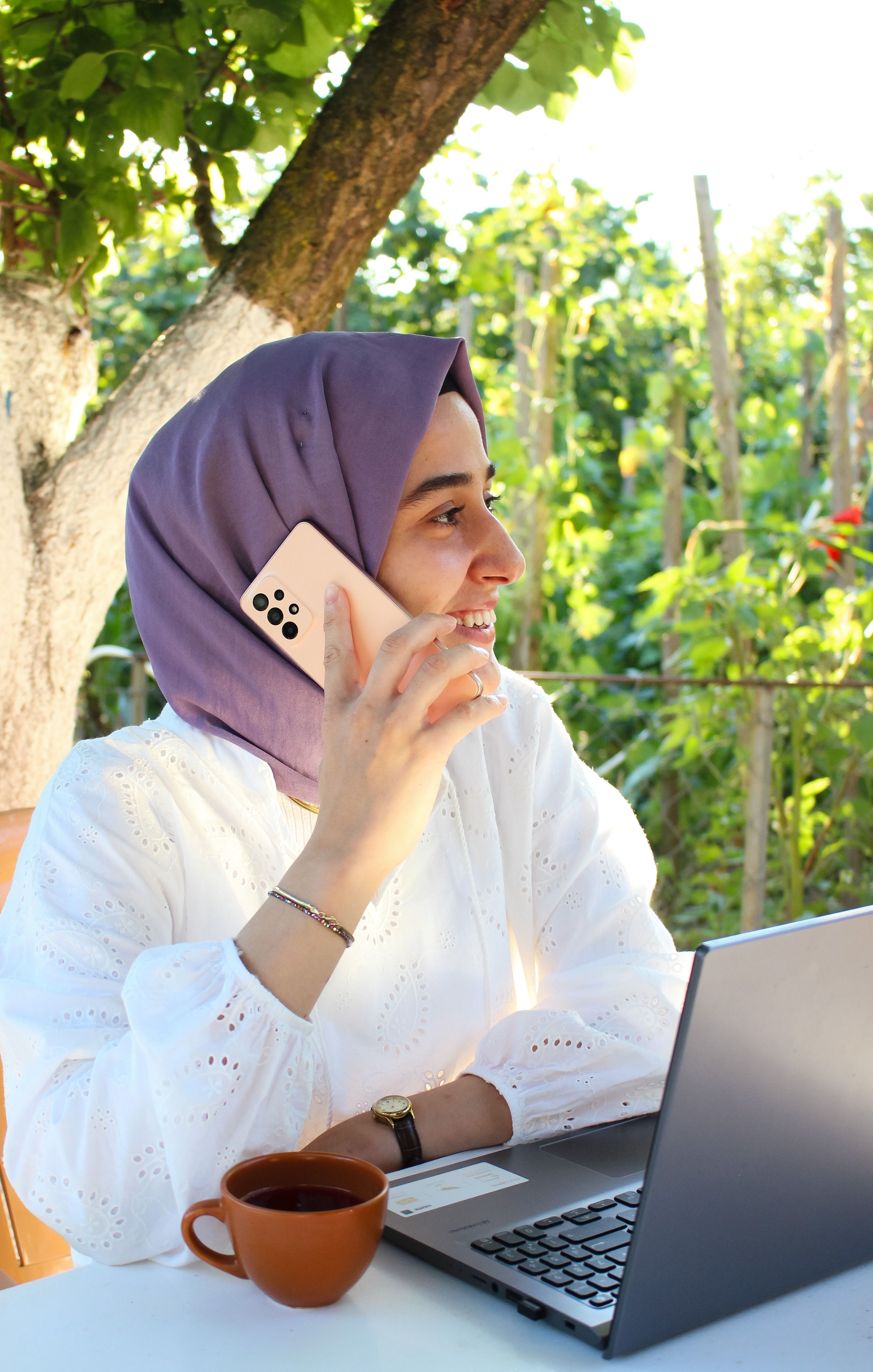 Smiling woman in hijab talking on phone while working on laptop outside.