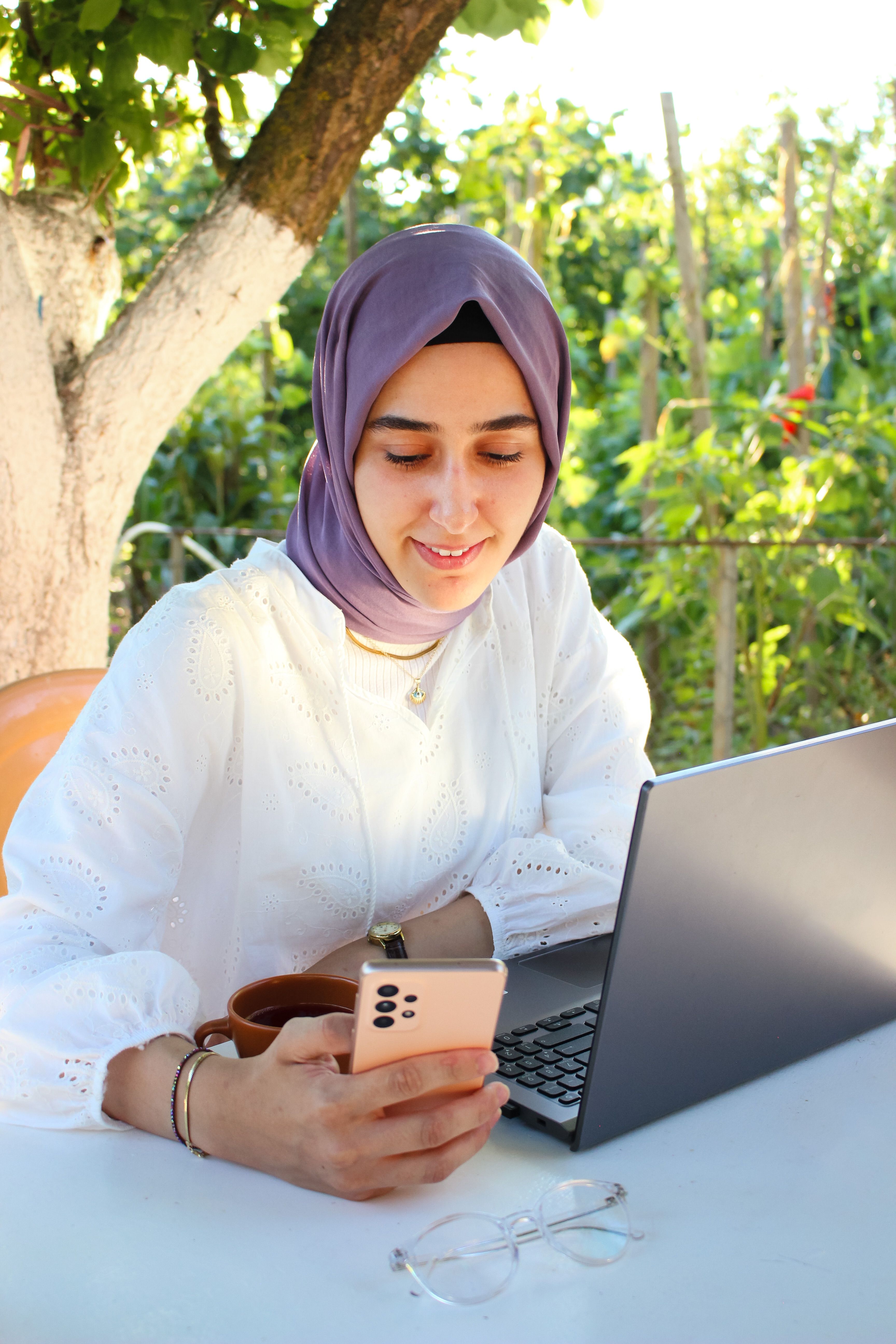 A woman in a hijab smiles as she uses her smartphone with a laptop on a sunny day.