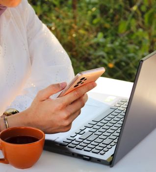 Close-up of a woman using a smartphone with a laptop and coffee cup outside.
