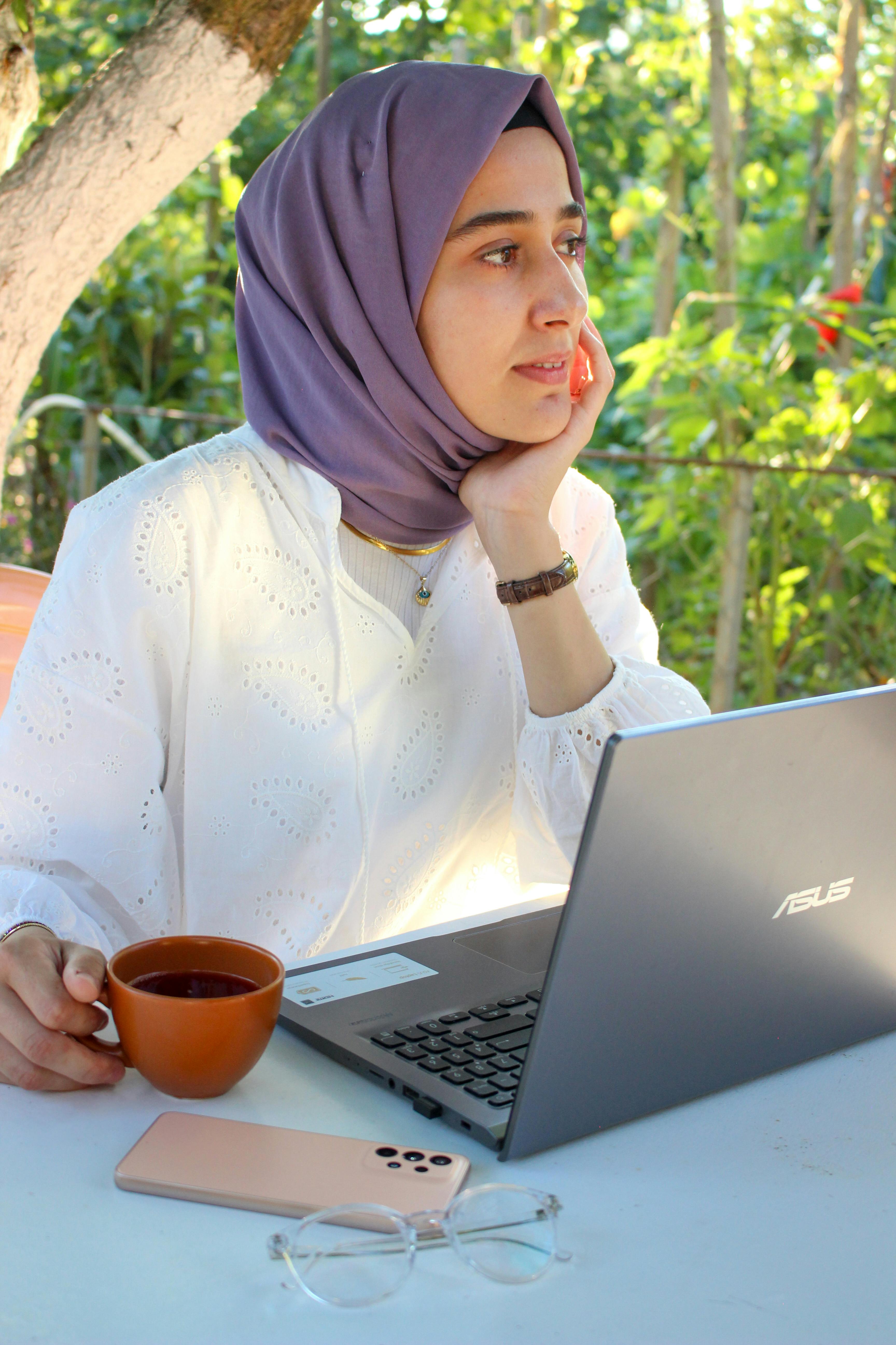 Woman in hijab working on a laptop outdoors, sipping coffee and holding a smartphone.