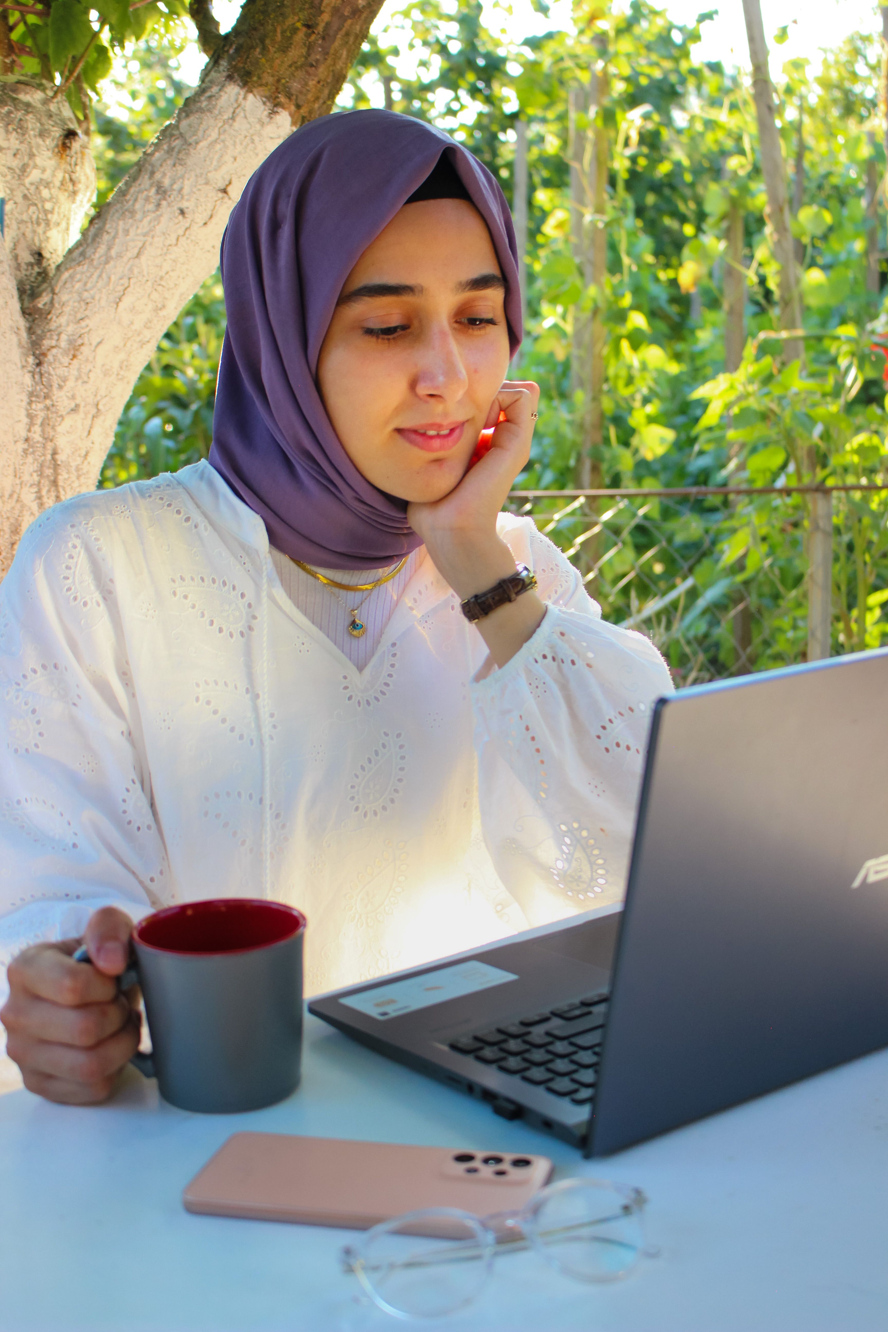 A woman in a hijab works outdoors on a laptop, holding a mug, amidst greenery.