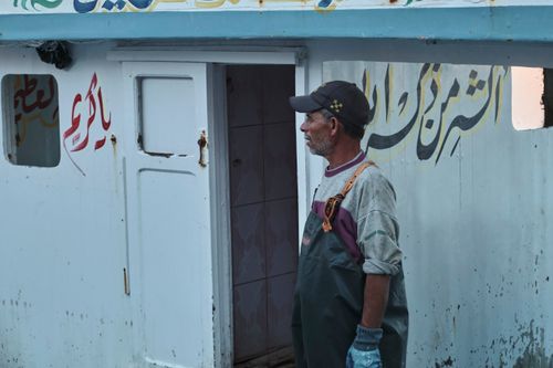 Free A man wearing work gear stands by a door with Arabic script, capturing a moment of daily life. Stock Photo