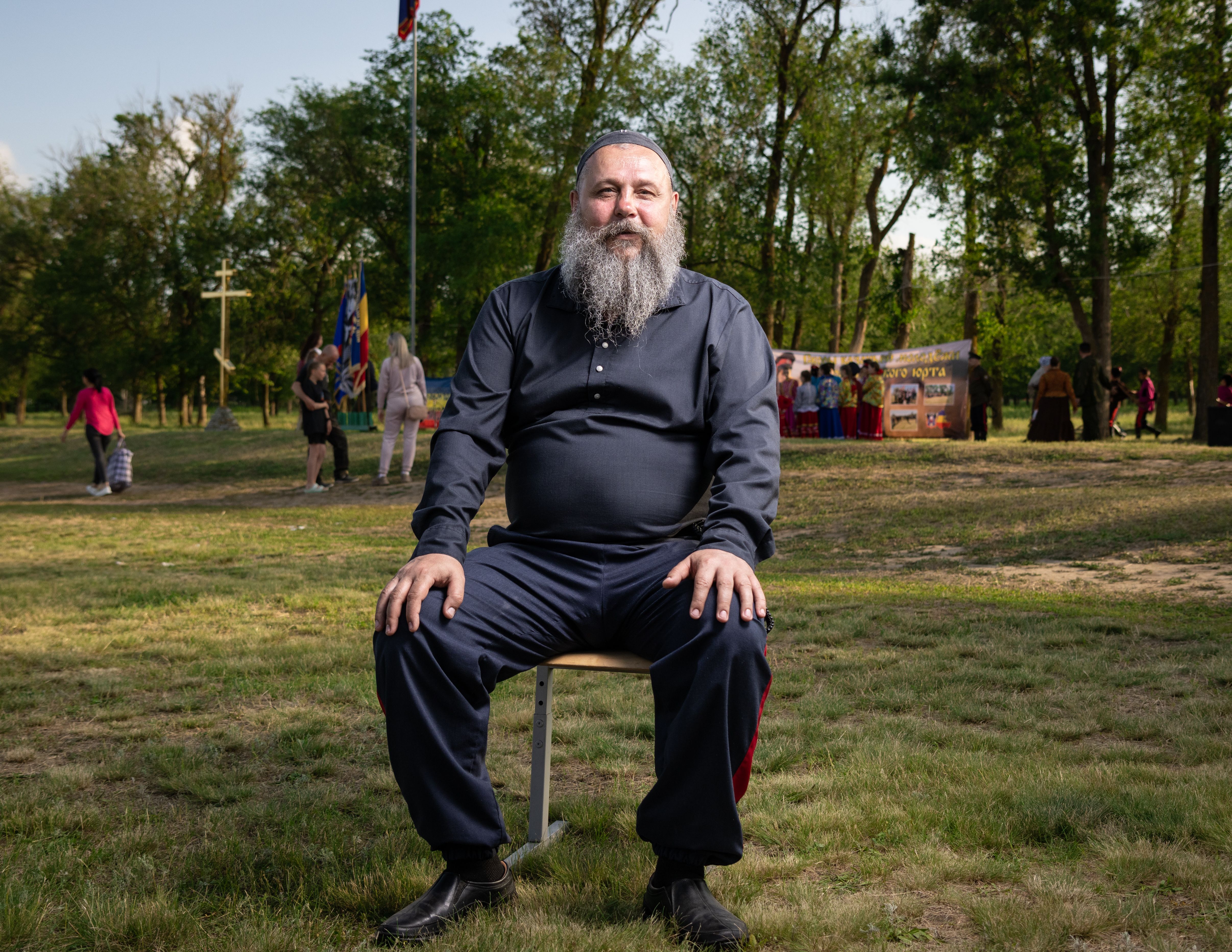 A man with a grey beard sits on a stool at an outdoor festival surrounded by trees.