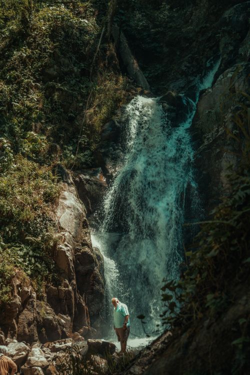 Free A senior man stands near a cascading waterfall in a lush, natural setting, conveying adventure and tranquility. Stock Photo