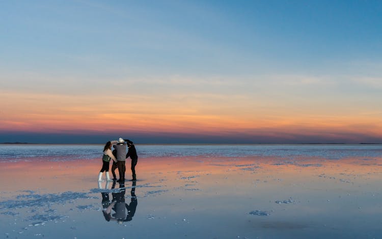 View Of A Group Of People Standing On A Beach At Sunset