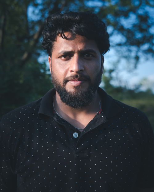Free A serious man with a beard stands outdoors in a sunlit Srinagar park surrounded by trees. Stock Photo