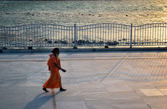 A Hindu monk in orange robes walks by the Ganges in Rishikesh, India, under soft evening sunlight.