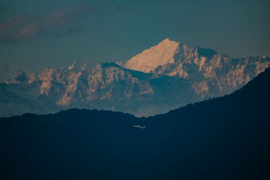 A solitary airplane flying below the majestic snowcapped mountain range during twilight, offering a serene landscape view.