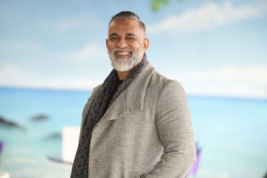 A smiling man in a grey coat posing with a beach backdrop, exuding warmth and confidence.