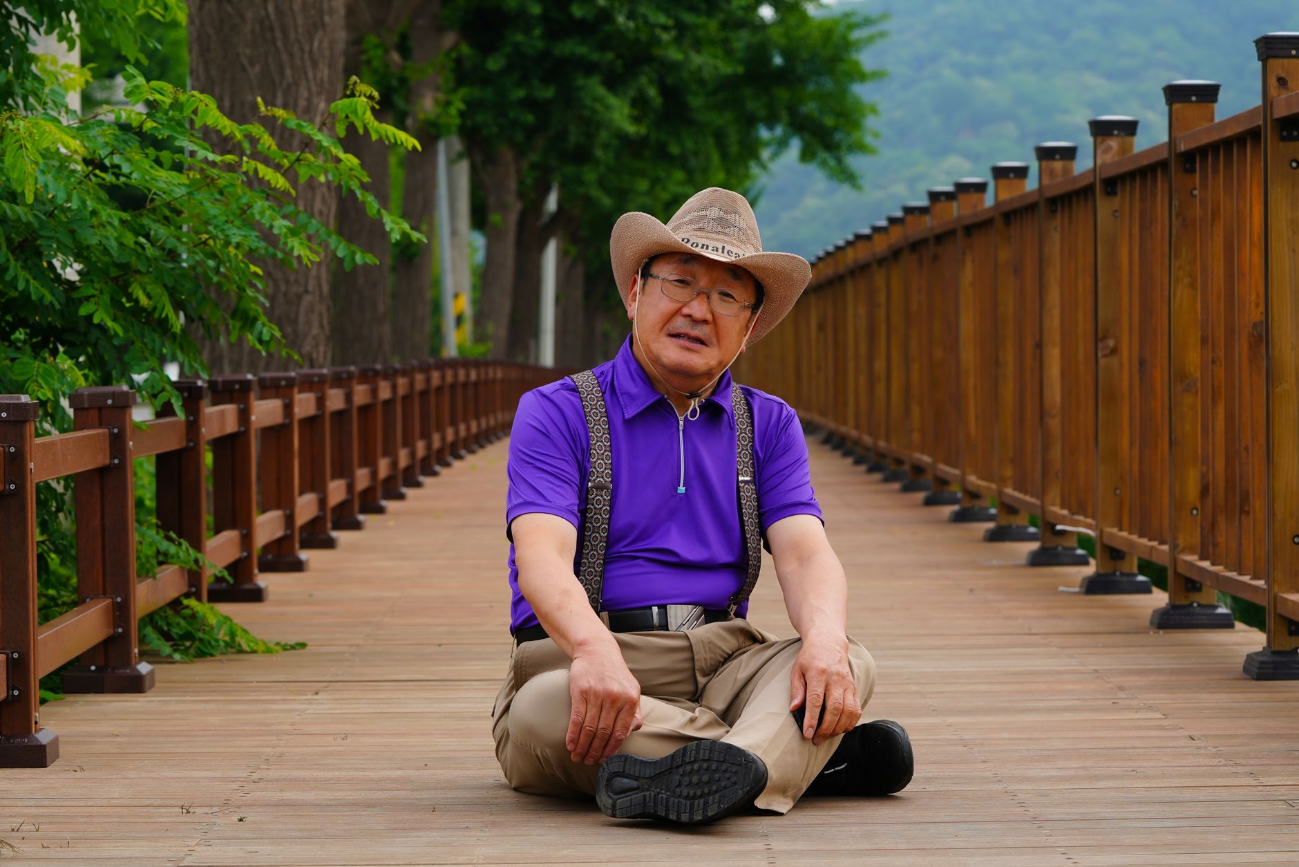 Senior man in a cowboy hat sitting cross-legged on a wooden footbridge surrounded by greenery.
