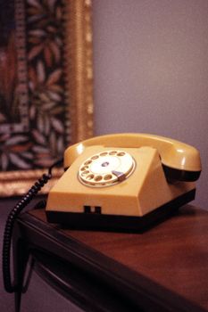 A vintage yellow rotary dial telephone on a wooden table with a framed painting in the background.