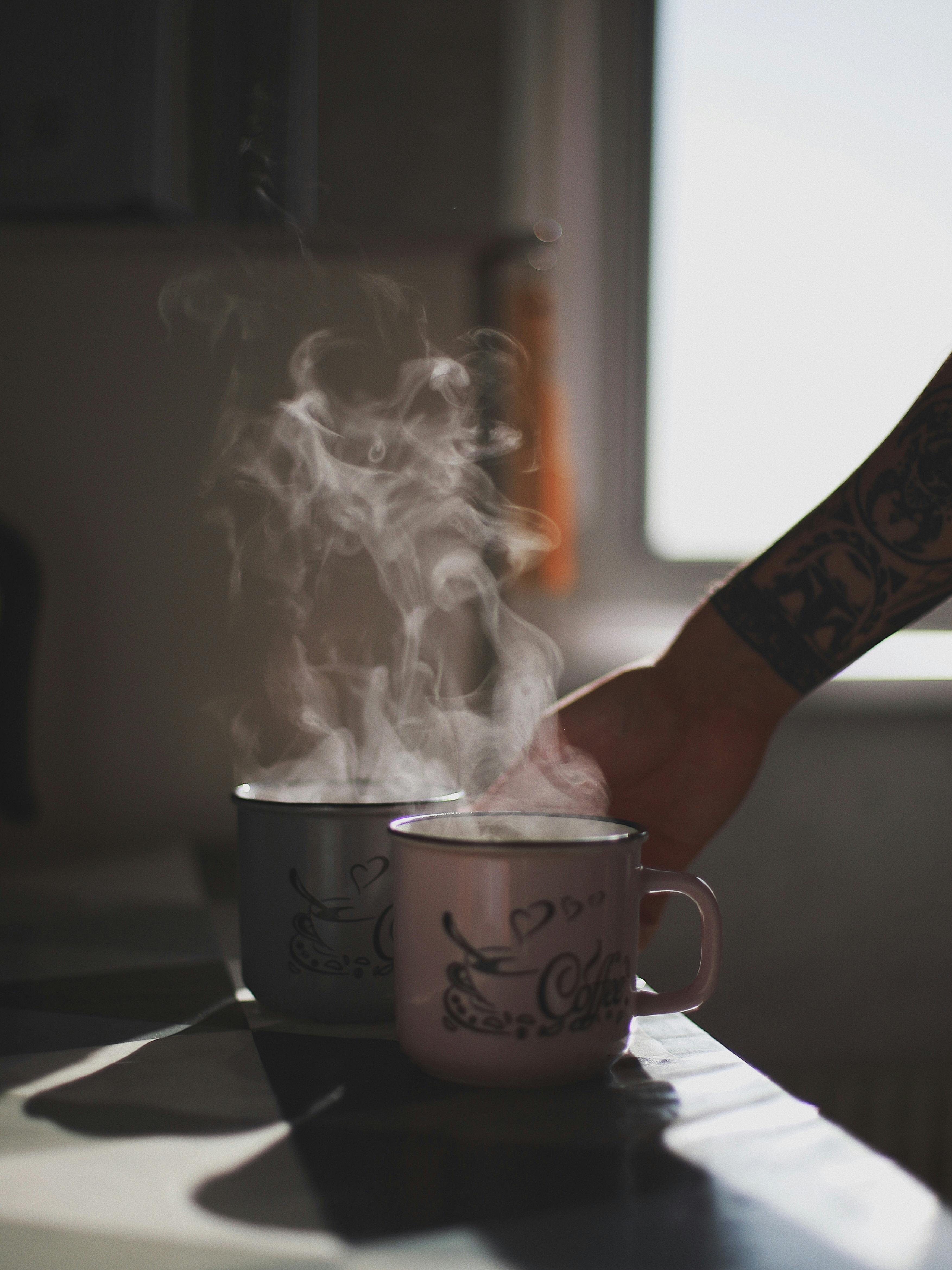 A tattooed hand reaches for a steaming mug of coffee indoors with soft lighting.