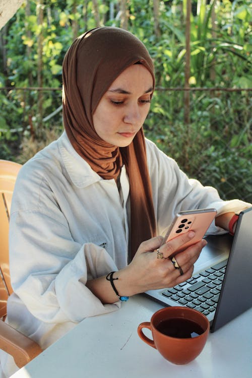 Portrait of a Young Woman Wearing a Hijab Using a Smart Phone in front of a Laptop
