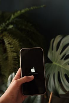 Close-up of a hand holding a smartphone with an Apple logo against a plant backdrop.