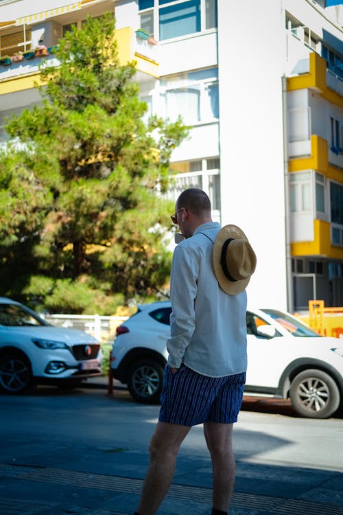 Man in Shorts, White Shirt and a Hat Standing on a Sidewalk in City 