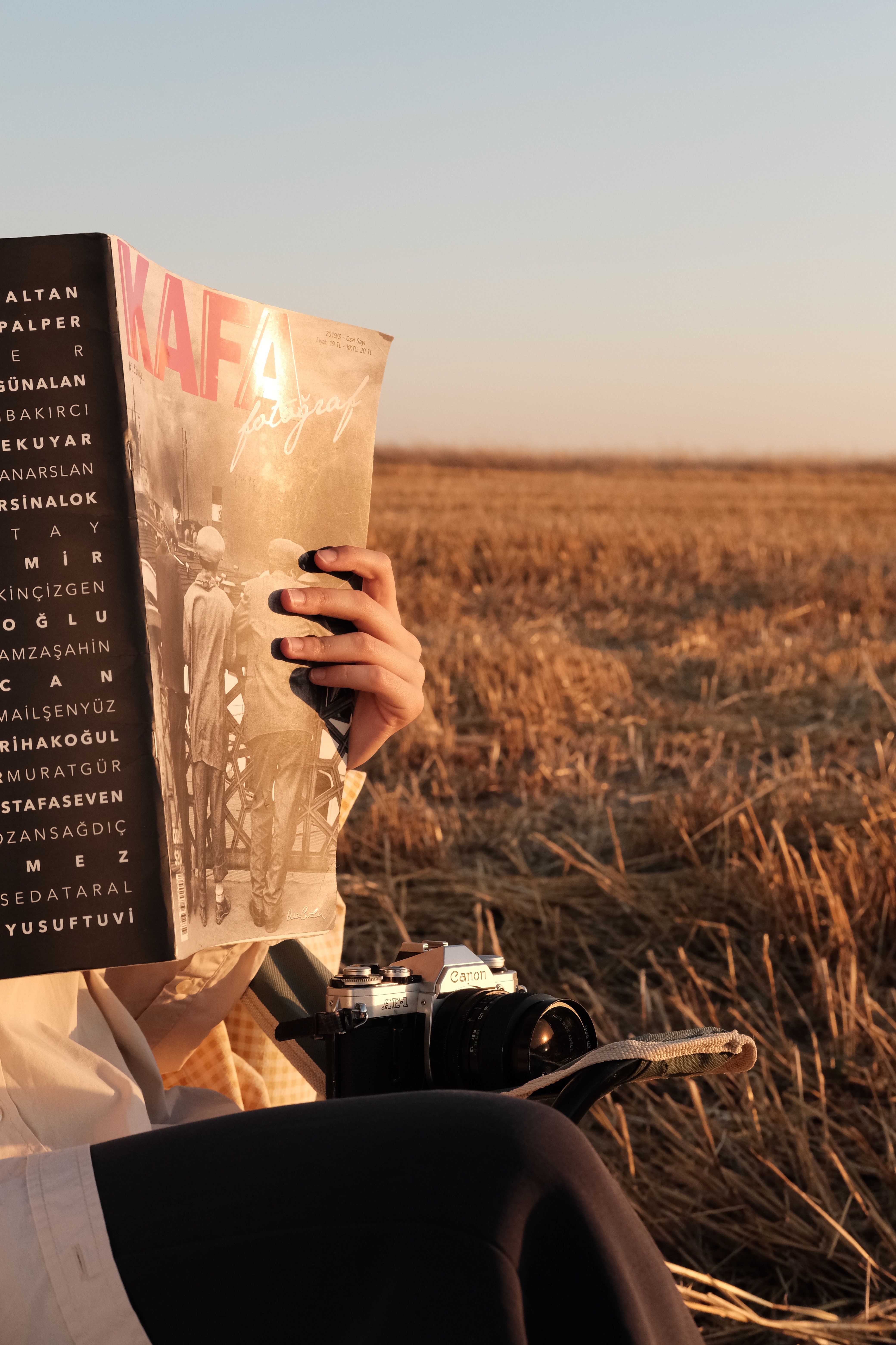 A person sits in a rural field at dusk, reading a magazine, capturing a moment of relaxation.