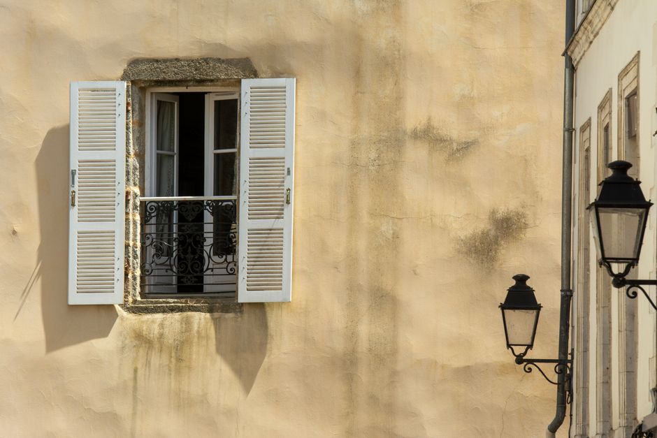 A rustic window with white shutters and ornate ironwork, casting shadows on a sunny day.