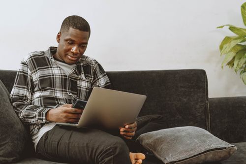Free Man using laptop and smartphone while sitting on a sofa in a cozy living room setting. Stock Photo