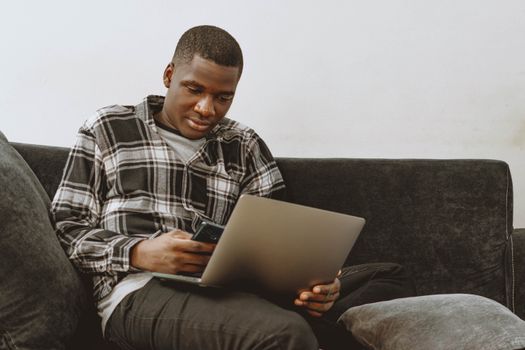 Young adult using laptop and smartphone on a sofa, focusing on remote work technology.