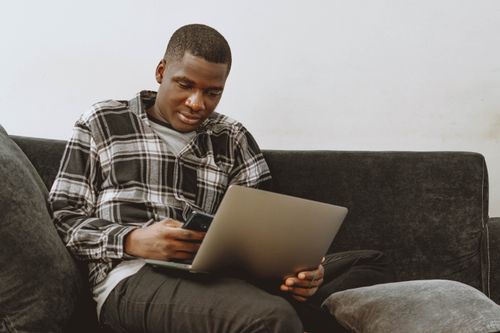 Free Young adult using laptop and smartphone on a sofa, focusing on remote work technology. Stock Photo