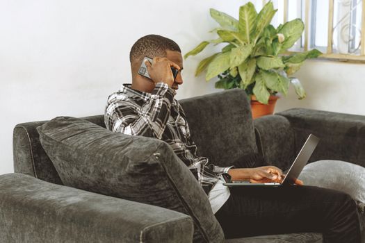 A young man multitasking with a smartphone and laptop on a sofa, illustrating remote work from home.