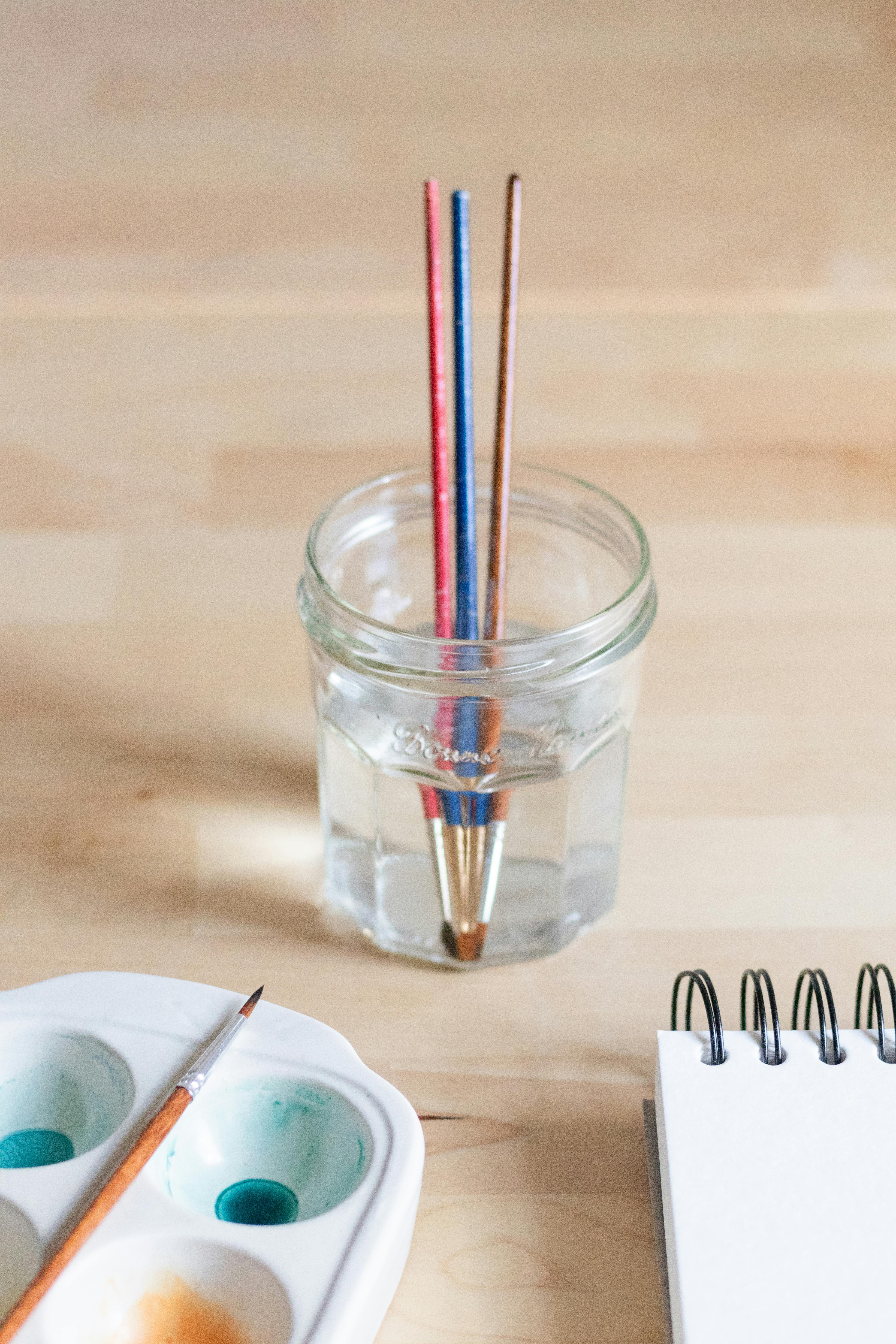 Glass jar with paintbrushes, watercolor paints, and notebook on a wooden table.