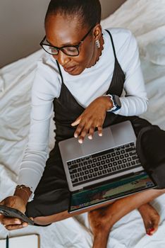 African woman multitasking with a laptop and smartphone on bed, showcasing modern technology.