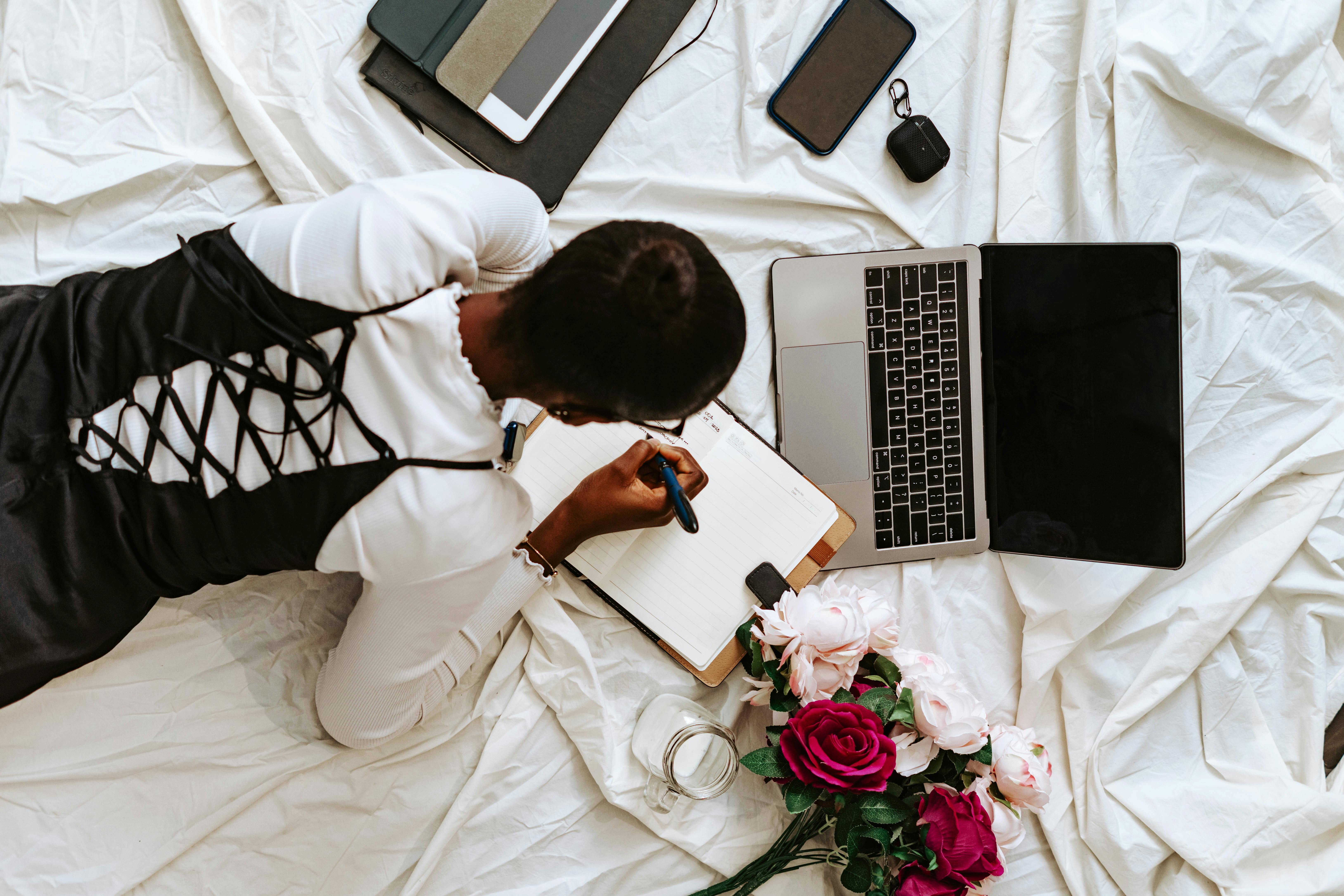 Free A young woman lies on a bed writing in a notebook beside a laptop and flowers, creating a serene workspace. Stock Photo