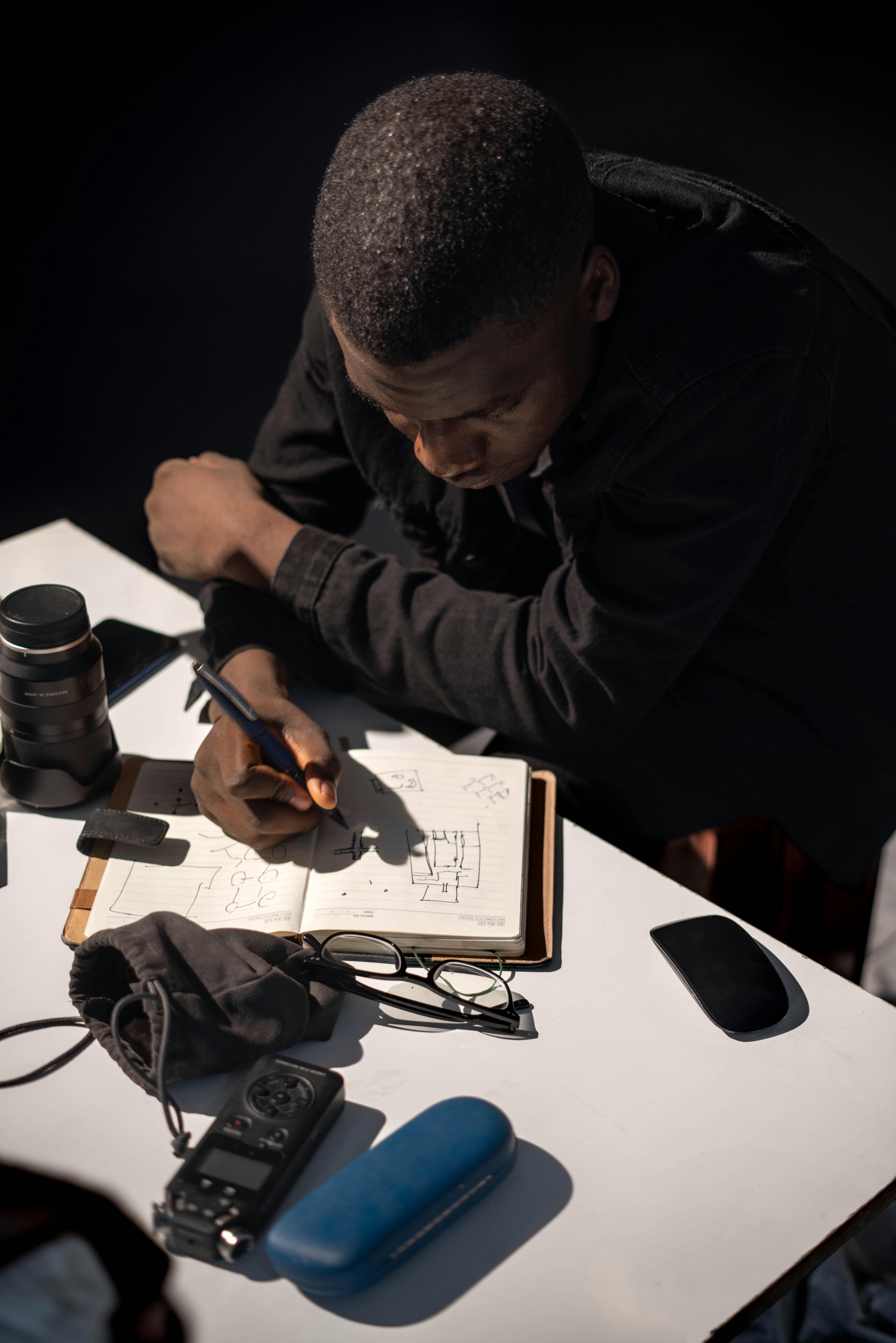 Free A man intensely sketching designs in a notebook, surrounded by tech gadgets, indoors. Stock Photo