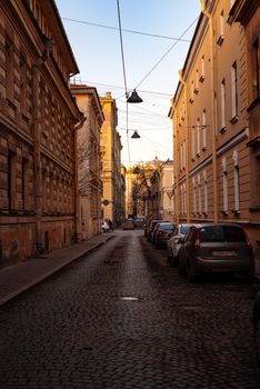 Narrow cobblestone street lined with buildings and parked cars, capturing urban tranquility at sunset.