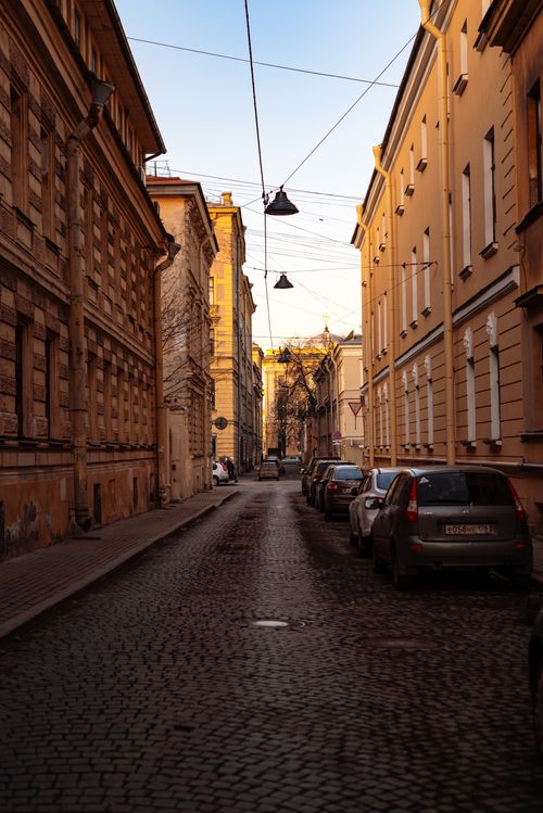 Free Narrow cobblestone street lined with buildings and parked cars, capturing urban tranquility at sunset. Stock Photo