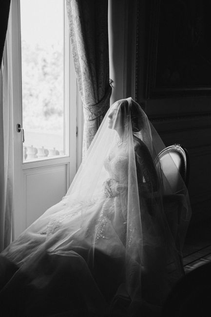 A stunning bride in a wedding dress sits gracefully by a window, enveloped in her veil, in a black and white portrait.