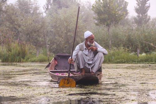 Man in White Kufi Cap Rowing in his Wooden Boat in a River