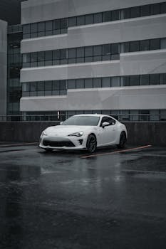 A white sports car parked in a city parking lot on a rainy day, showcasing its modern design.