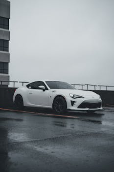 A sleek white sports car is parked in an urban setting with wet pavement reflecting a rainy day atmosphere.
