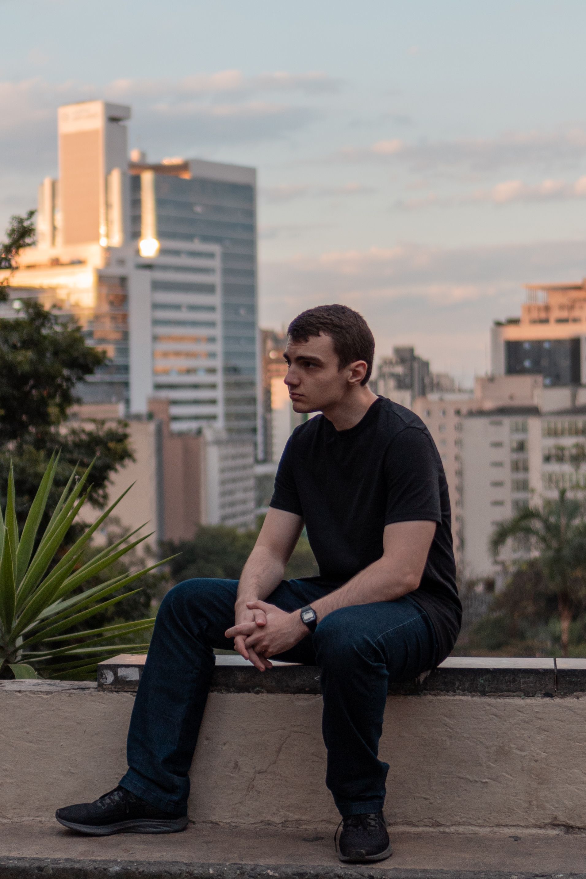A man sits pensively on a wall, overlooking a city's skyline at sunset. Urban lifestyle.