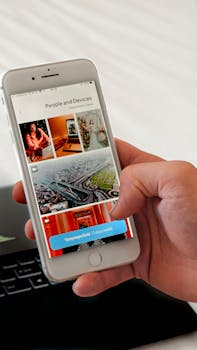 Hand browsing through photos on a smartphone with a laptop in the background indoors.