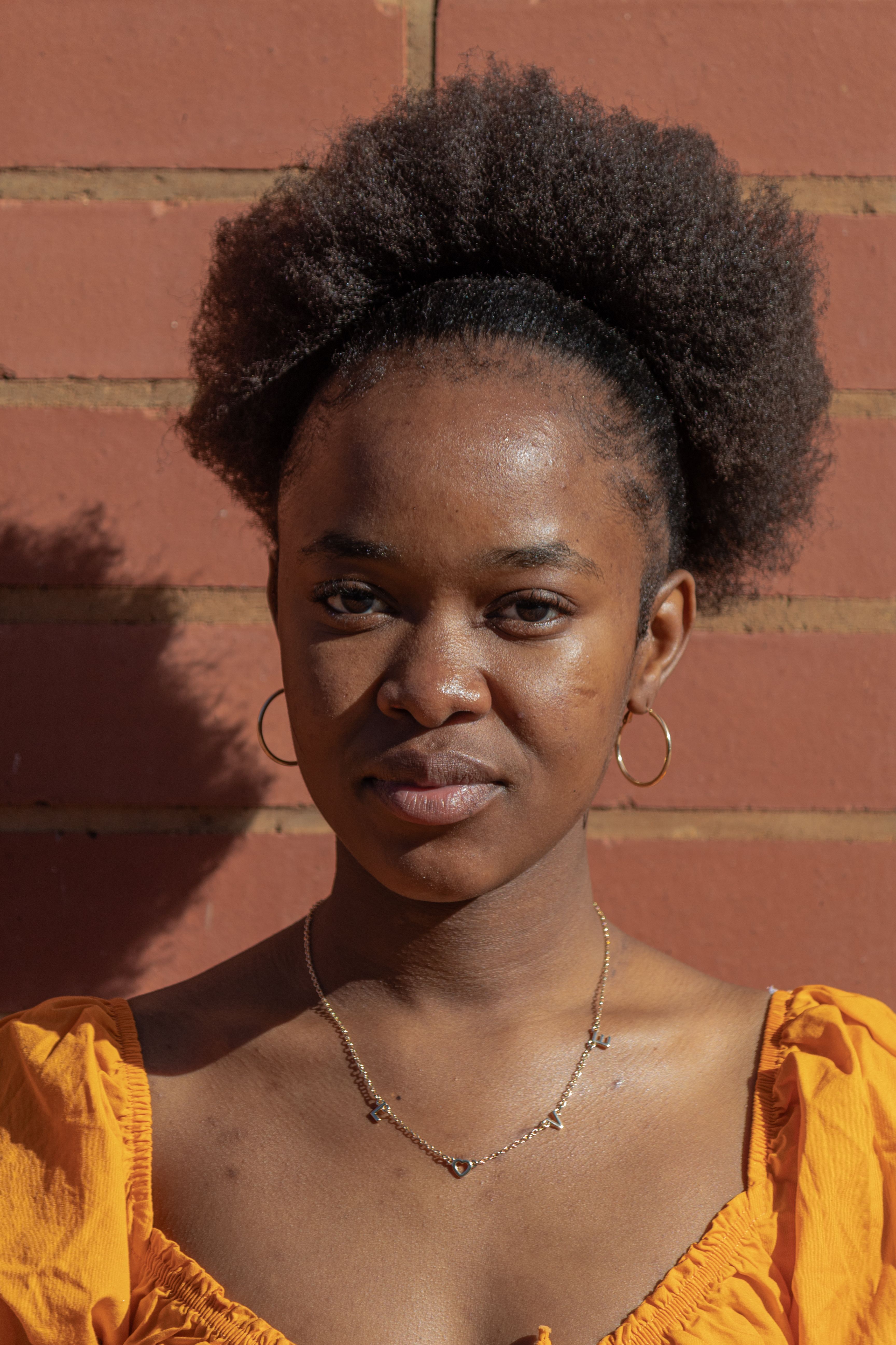 Close-up portrait of a young woman against a brick wall, highlighted by warm sunlight.