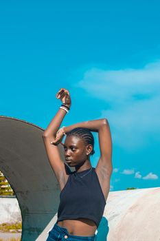 A fashionable young black woman poses outdoors in the summer sun, showcasing modern style with a blue sky backdrop.