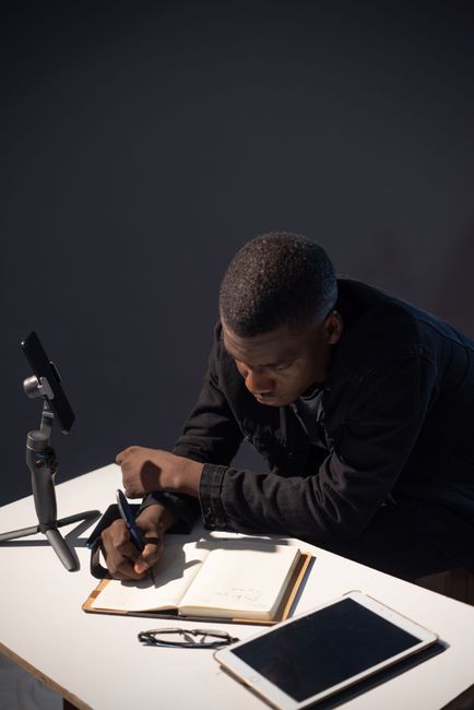 Young man using tech gadgets and writing notes, focusing in a dark room setting. Young man using tech gadgets and writing notes, focusing in a dark room setting.