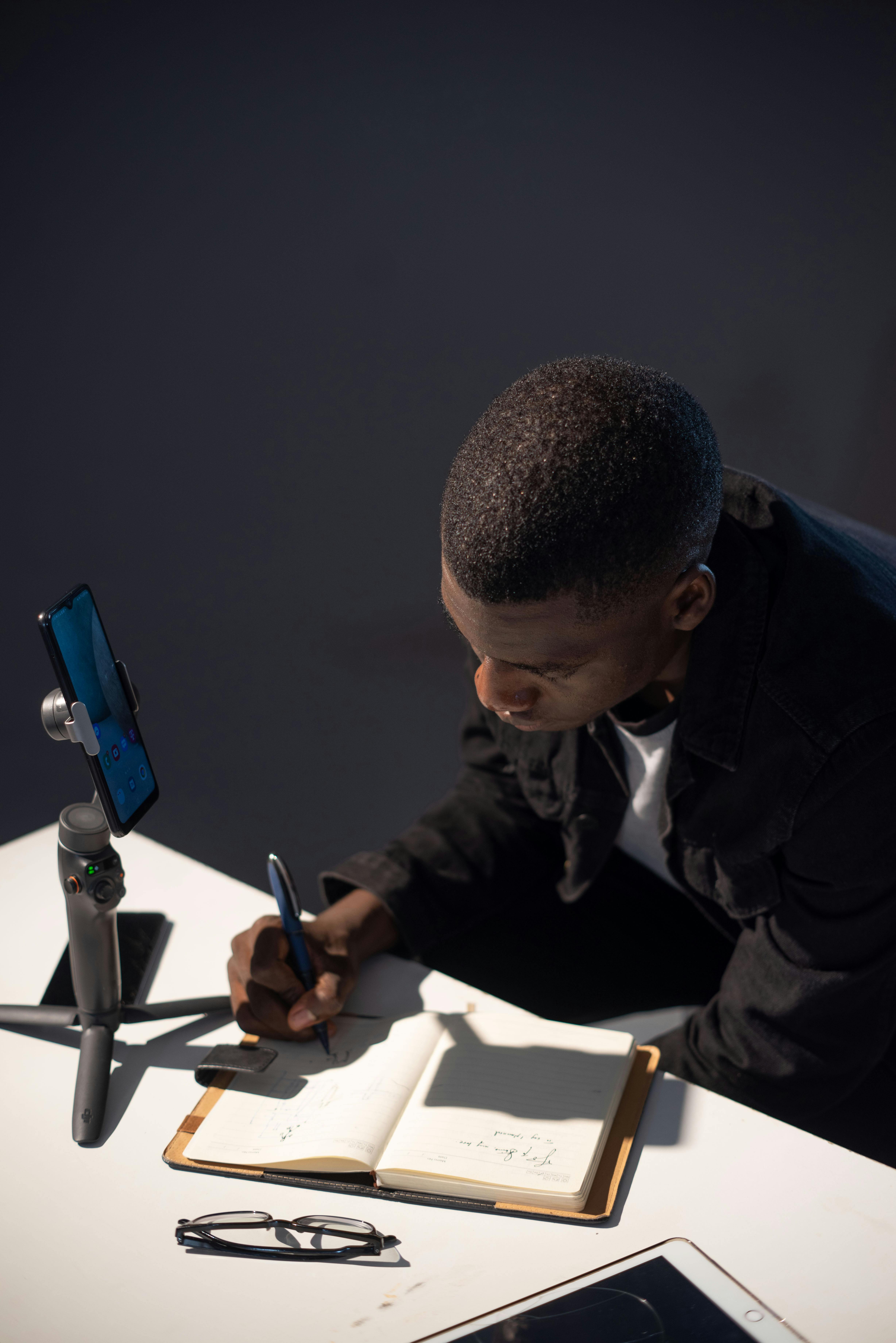 Free A young man concentrates on writing in a notebook while using a smartphone gimbal, symbolizing modern remote work. Stock Photo