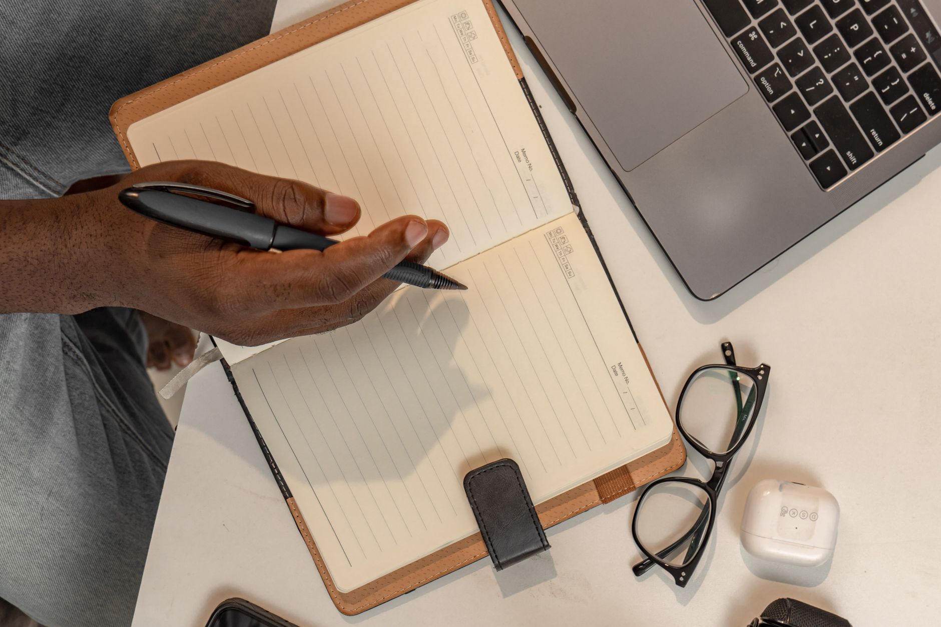 Man writing in a notebook at desk with laptop and eyeglasses. Organized workspace concept.