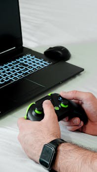 Close-up of hands holding a gaming controller near a laptop with LED keyboard.