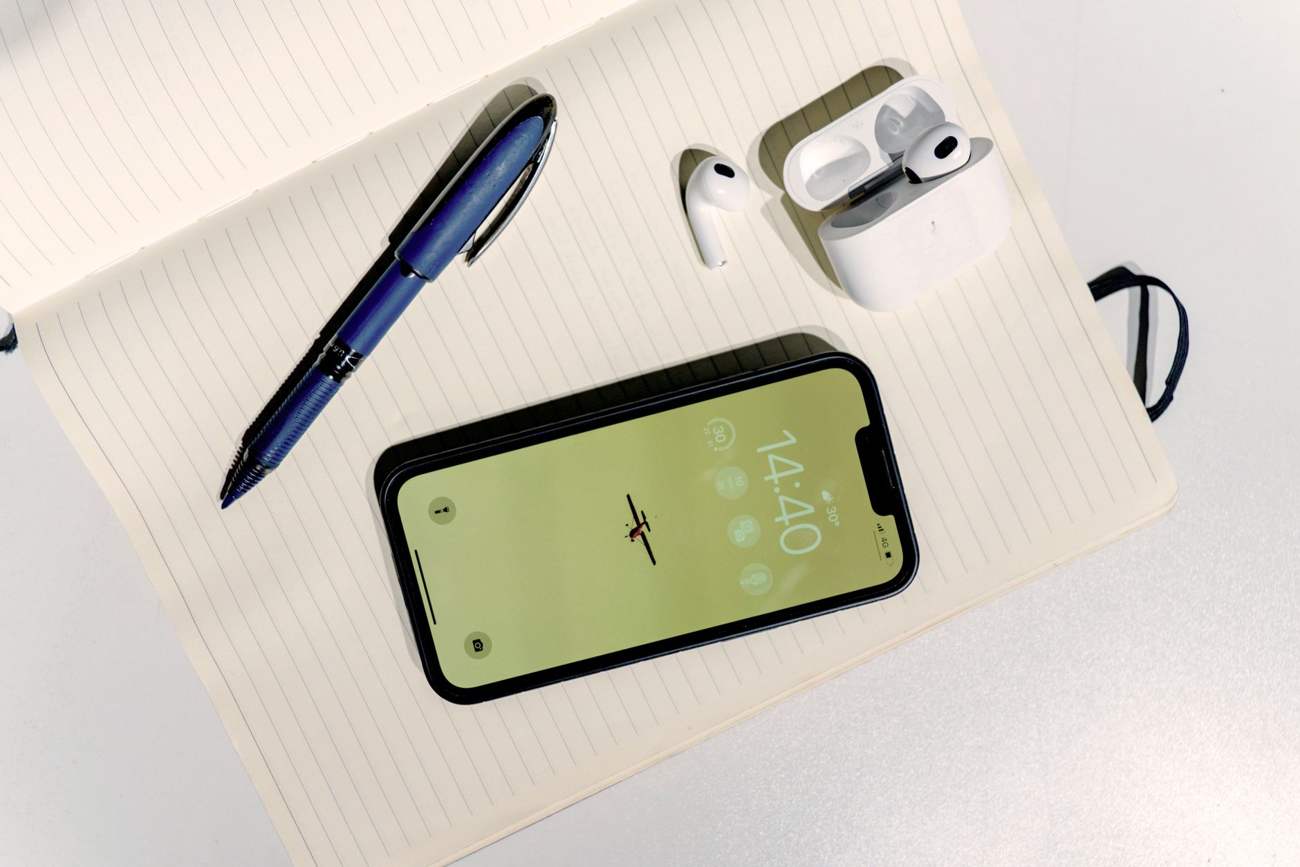 A minimalist flat lay featuring a smartphone, wireless earbuds, pen, and notebook on a light desk.
